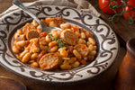 A close-up of a bowl of beans and sausage in a tomato-based sauce, garnished with fresh thyme, served on a patterned ceramic plate with a spoon.