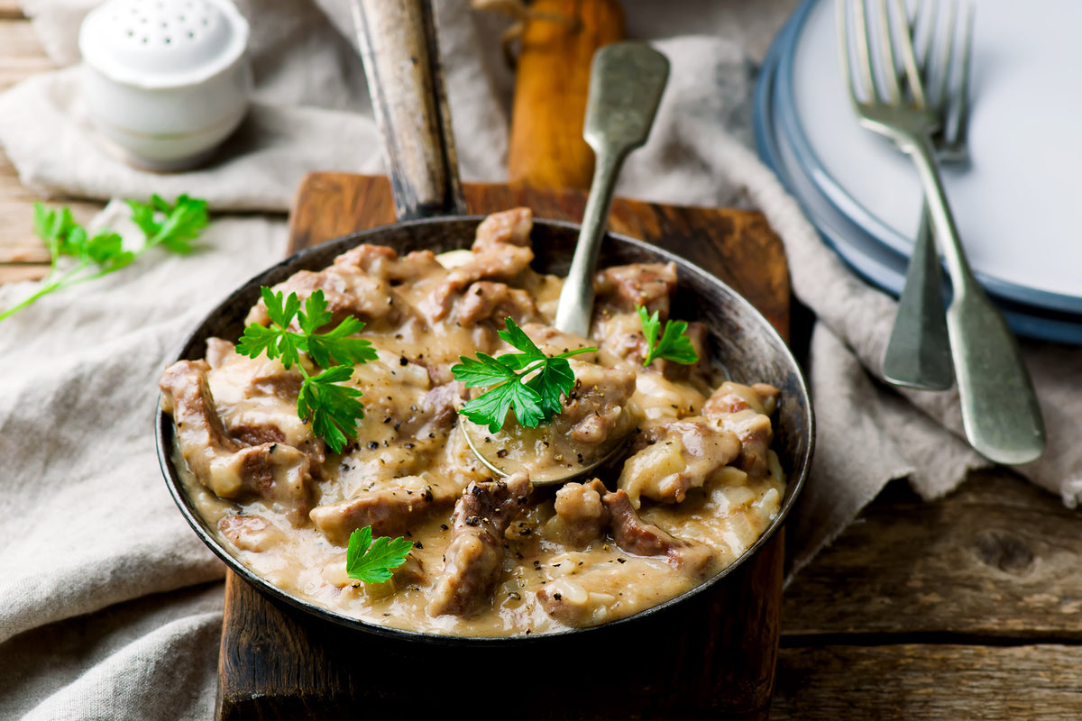 A cast iron skillet filled with creamy dijon sauce over beef, garnished with fresh parsley, set on a wooden board with a spoon and fork in a rustic dining setting.