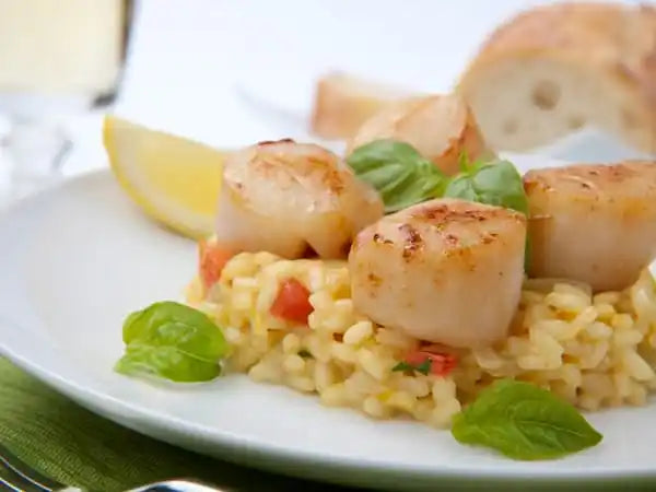 Scallops on a bed of pasta with a lemon wedge and bread on a white plate.